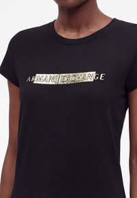 T-shirt noir à manches courtes avec texte doré métallique brillant "Armani Exchange" sur la poitrine, porté par une personne à la peau foncée.