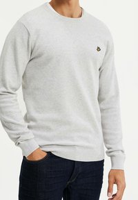 Grå texturerad sweatshirt med rund halsringning, ribbade muddar och en liten broderad logga på bröstet, kombinerad med mörka jeans.