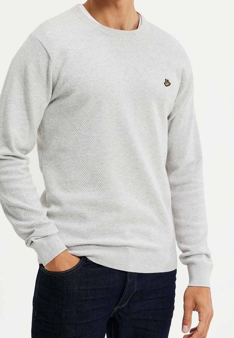 Grå texturerad sweatshirt med rund halsringning, ribbade muddar och en liten broderad logga på bröstet, kombinerad med mörka jeans.