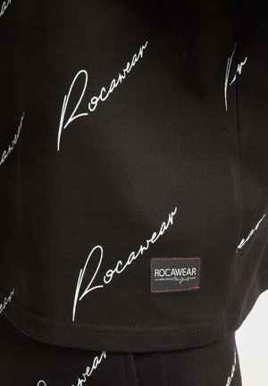 Melnas audums ar atkārtotu baltu "Rocawear" logo kursīvā. Ietver nelielu taisnstūra logo uzlīmi ar sarkaniem diegiem.