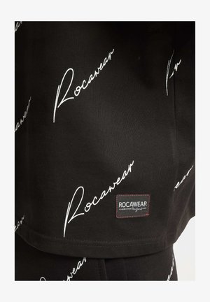 Material textil negru cu un logo alb "Rocawear" repetat, scris cu litere cursive. Include un mic eticheta logo rectangulară cu cusături roșii.