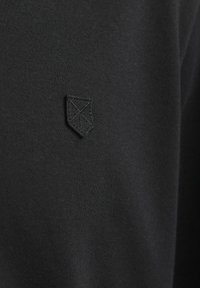 Fekete textil, amelyen egy kis fekete hímzett pajzs logó található geometrikus részletezéssel egy sima, egyenletes szövetfelületen.