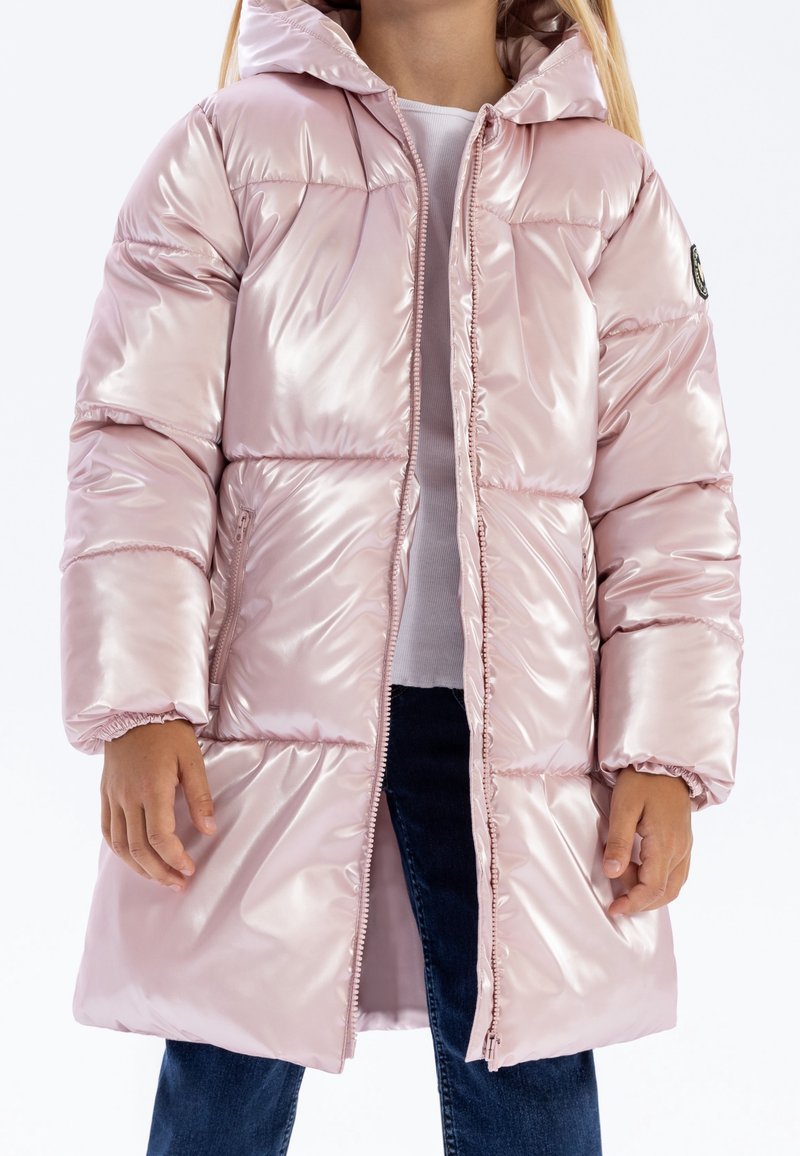 Enfant portant un long manteau matelassé rose pâle brillant avec capuche, chemise blanche et jean bleu foncé, debout sur fond blanc.