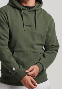 Olivgrön hoodie med ficka fram, dragsko-hets och broderad logotyp. Ribbad mudd och nederkant, strukturerad tyg.