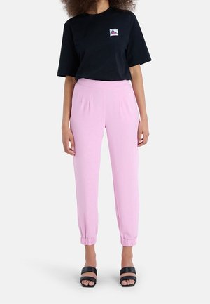 Kvinde iført en sort t-shirt med korte ærmer og et lille logo på brystet, lyserøde joggerbukser med høj talje og sorte åbne sandaler.