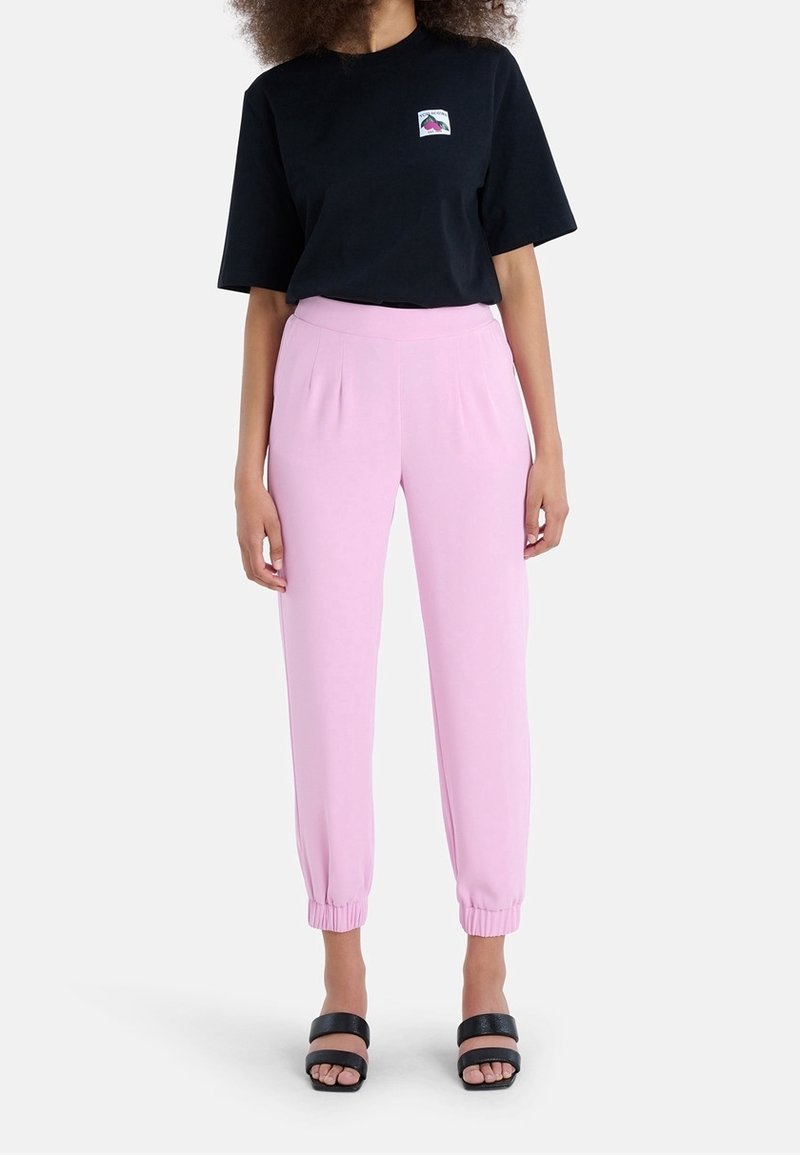 Frau trägt ein schwarzes T-Shirt mit kurzen Ärmeln und kleinem Brustlogo, pinke Jogginghosen mit hohem Bund und schwarze Sandalen mit offenem Zehenbereich.