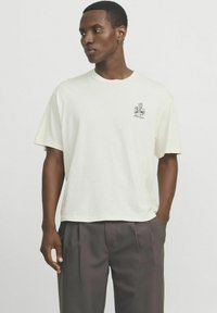 T-shirt en coton crème à coupe décontractée, manches courtes, avec un petit motif floral noir sur la poitrine. Porté avec un pantalon gris plissé.