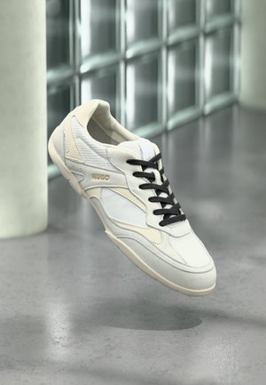 Biały i beżowy sneaker z czarnymi sznurowadłami i logo „HUGO”, unoszący się nad błyszczącą szarą podłogą, na tle szklanej ściany z bloków.