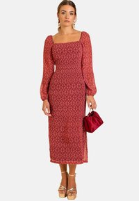 Rotes gemustertes Langarmkleid mit quadratischem Ausschnitt und taillierter Taille, kombiniert mit einer kleinen roten Tasche und goldenen Plateauschuhen.