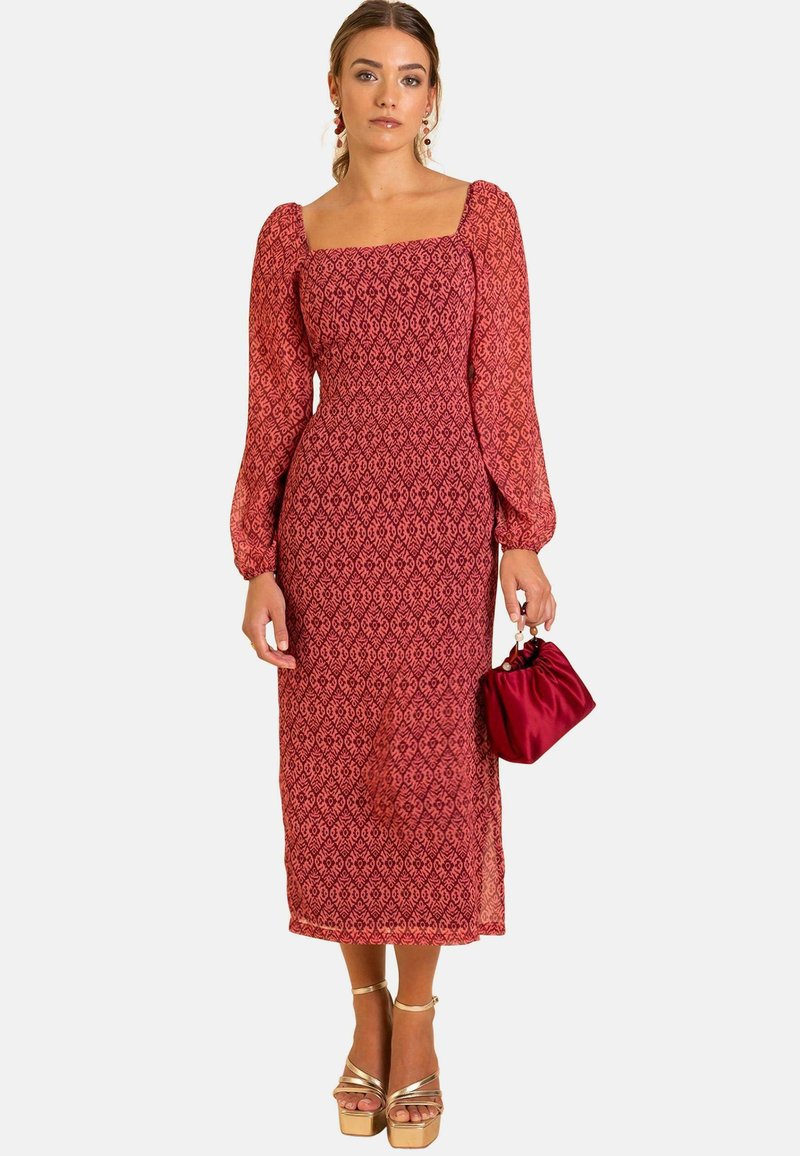 Rotes gemustertes Langarmkleid mit quadratischem Ausschnitt und taillierter Taille, kombiniert mit einer kleinen roten Tasche und goldenen Plateauschuhen.