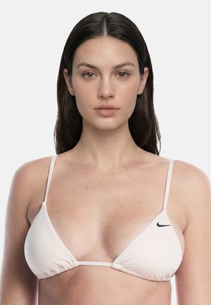 Joven mujer con largo cabello oscuro que lleva un bikini triangular blanco de Nike, mirando hacia adelante sobre un fondo liso.
