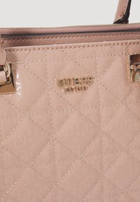 Jasnoróżowa torebka z syntetycznego materiału o wypukłym wzorze logo i złotych detalach. Zamek błyskawiczny z dopasowanymi akcentami na pasku.