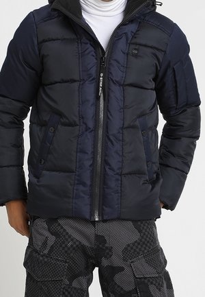 Mann trägt eine marineblaue gesteppte Jacke mit Fronttaschen und Tarn-Cargohosen, teilweise offen über einem weißen Rollkragenpullover.