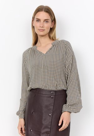 Woman with straight blonde hair wears a loose checked blouse and dark brown buttoned leather skirt, standing against a white background.