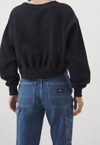 Svart kort sweatshirt med ribbat nederkant och muddar, ihopparad med blå denimbyxor som har två bakfickor och en logotypbricka.