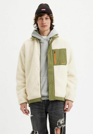 Chaqueta de forro polar con tela verde oliva, cuerpo texturizado en crema, bolsillo con cremallera naranja, combinada con una sudadera gris y jeans negros desgastados.