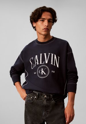 Mladý muž s kučeravými vlasmi nosí čiernu mikinu Calvin Klein a tmavé džínsy, stojí s jednou rukou v vrecku na sivom pozadí.