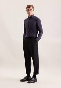 Camicia blu navy scura con bottoni, pantaloni neri plissettati e scarpe di pelle nere. Tessuto liscio con vestibilità sartoriale e dettagli di design sottili.