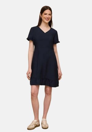 Ung kvinde stående i en kort sort kjole med flæser på ærmerne og kanten, kombineret med beige sneakers, smilende mod en ensfarvet baggrund.