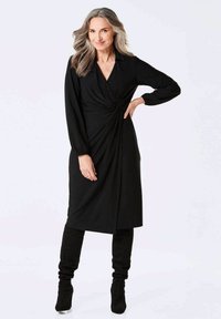 Schwarzes Wickelkleid mit langen Ärmeln, gerafftem Seitendetail und einem Kragen. Model trägt knielange schwarze Stiefel, weicher Stoff mit glatter Textur.