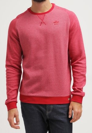 Homme portant un pull rouge à manches longues avec une texture, des poignets, un ourlet et un col rond côtelés, associé à un jean foncé.