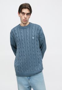 Μπλε πλεγμένο πουλόβερ με σχέδιο κοτσίδα, στρογγυλή λαιμόκοψη και ριμπ μανσέτες και τελείωμα. Φορεμένο με ανοιχτόχρωμο τζιν, παρουσιάζοντας άνετη εφαρμογή.