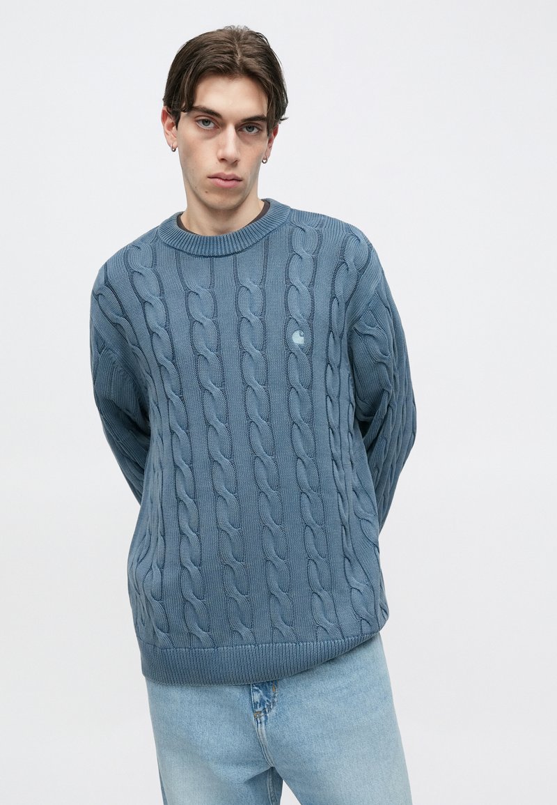 Blauwe gebreide trui met een kabelpatroon, ronde hals en geribbelde boorden en zoom. Draagt lichtblauwe broek, met een ontspannen pasvorm.