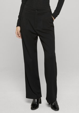 Kobieta ubrana w czarne szerokie spodnie z wysokim stanem i plisami z przodu, czarną bluzkę z długim rękawem oraz czarne botki na obcasie, stojąca z rękami w kieszeniach.