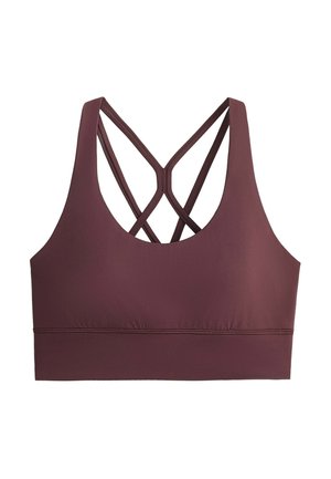 Femme portant un soutien-gorge de sport grenat et un pantalon de survêtement bleu marine, tenant un sac de sport beige, debout devant un fond gris.