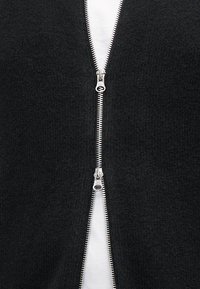 Sort zip-up sweater med en struktureret strikket overflade, der har en sølvlynlås ned langs midten og en hvid skjorte under.