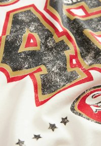 Tessuto bianco con stampa grafica rossa, nera e dorata. Presenta grandi numeri stilizzati e stelle in una texture invecchiata.