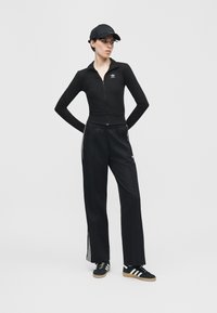 Vrouw in een zwart Adidas trainingspak met drie strepen, een zwarte pet en zwarte Adidas sneakers, staand met de handen achter de rug.