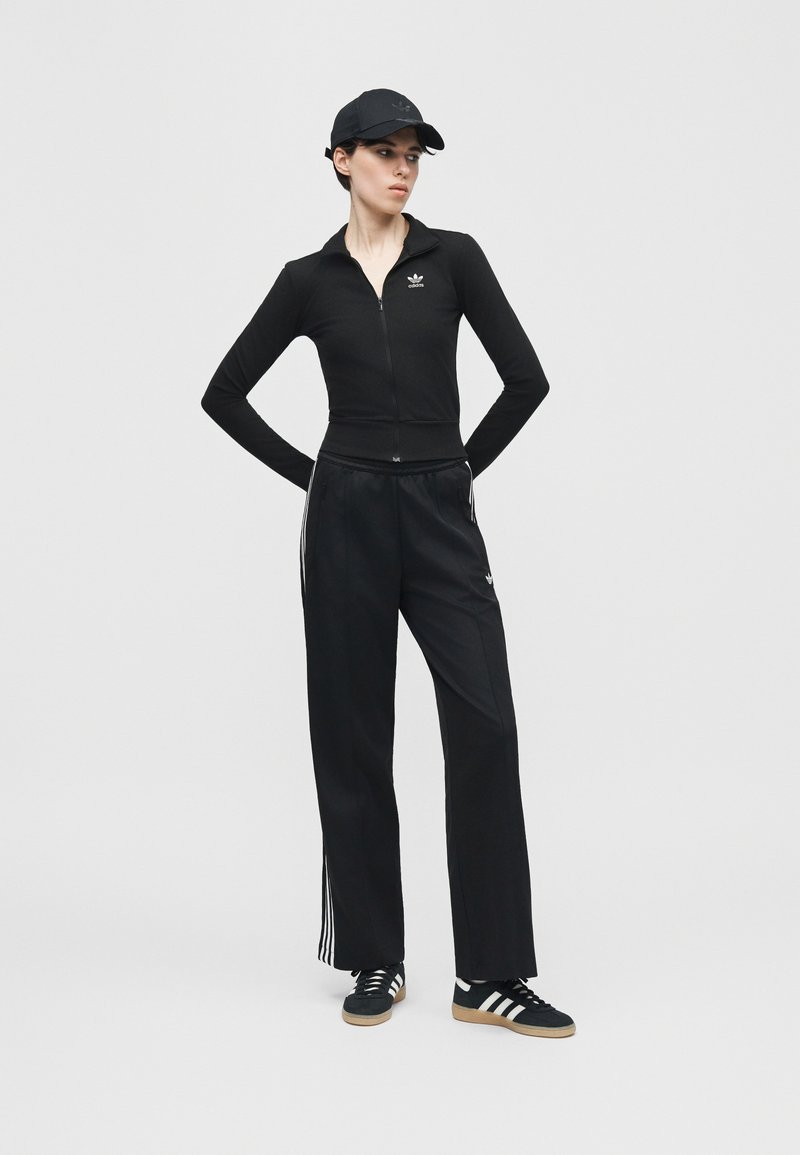 Femme portant un survêtement Adidas noir avec trois bandes, une casquette noire et des baskets Adidas noires, debout les mains derrière le dos.