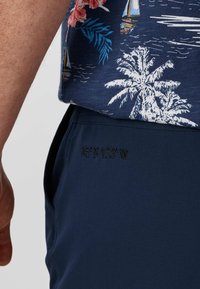 Homme portant un pantalon bleu marine avec des coordonnées brodées et une chemise bleu marine imprimée de palmiers blancs et de voiliers.