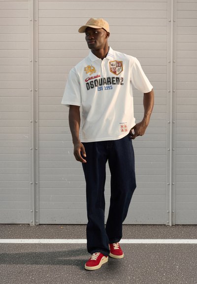 Polo blanc avec des motifs noirs audacieux et colorés, jean bleu marine et baskets rouges avec semelles crème. Casquette de baseball beige clair.
