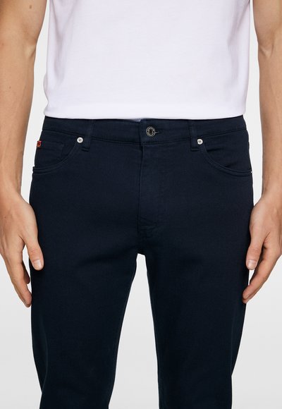 Tmavomodré džínsy slim fit s jedným gombíkom, prednými vreckami a malým červeným logom na zadnej strane.
