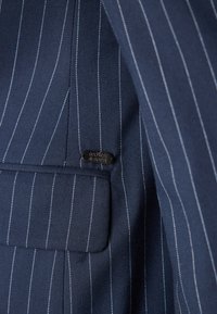 Navy blue pinstripe fabric with a smooth texture, featuring a pocket and a small metal tag embossed with "SCOTCH & SODA."