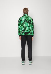 Giacca verde e nera con motivo, colletto alto, polsini neri, abbinata a pantaloni neri e scarpe da ginnastica multicolori.