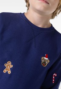 Marinblå sweatshirt med rund halsringning som har broderade pepparkaksgubbar, en mops med tomteluva och godiskäppar som accenter. Mjuk textur.