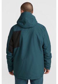 Chaqueta impermeable en color azul verdoso con un parche negro; cuenta con capucha y un corte ligeramente holgado, fabricada con una tela suave y duradera.