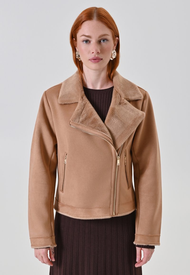 Donna con capelli rossi che indossa una giacca beige foderata in pelliccia sintetica sopra un vestito a coste scuro, in piedi contro uno sfondo semplice chiaro.