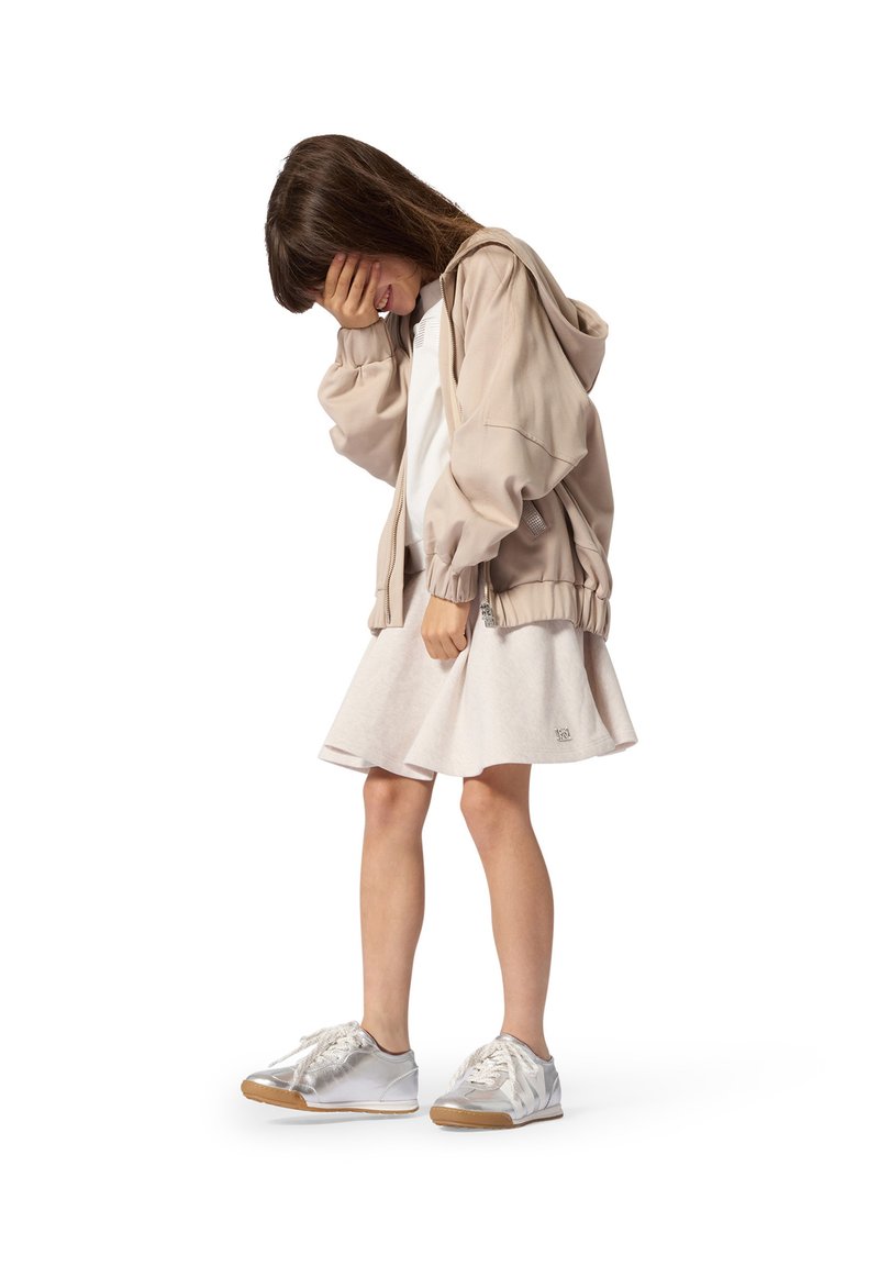 Jeune fille en veste beige et robe blanche, debout la tête baissée, couvrant son visage d'une main, portant des baskets blanches.
