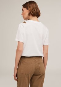 Camisa de algodón blanca de manga corta, con un acento texturizado dorado en el hombro. Combinada con unos pantalones marrones claros.