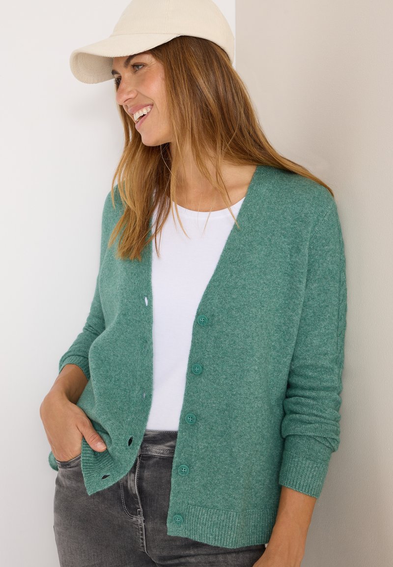 Grüne gestrickte Strickjacke mit V-Ausschnitt, Knopfleiste, gerippten Bündchen und Saum. Kombiniert mit einem weißen T-Shirt und grauen Jeans. Cremefarbener Hut.