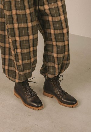 Rudi ir juodi batraiščiais įrišti batai su tvirtais padais, dėvimi su laisvais rudos ir žalios spalvų languotais kelnėmis, stovint ant šviesios grindų.