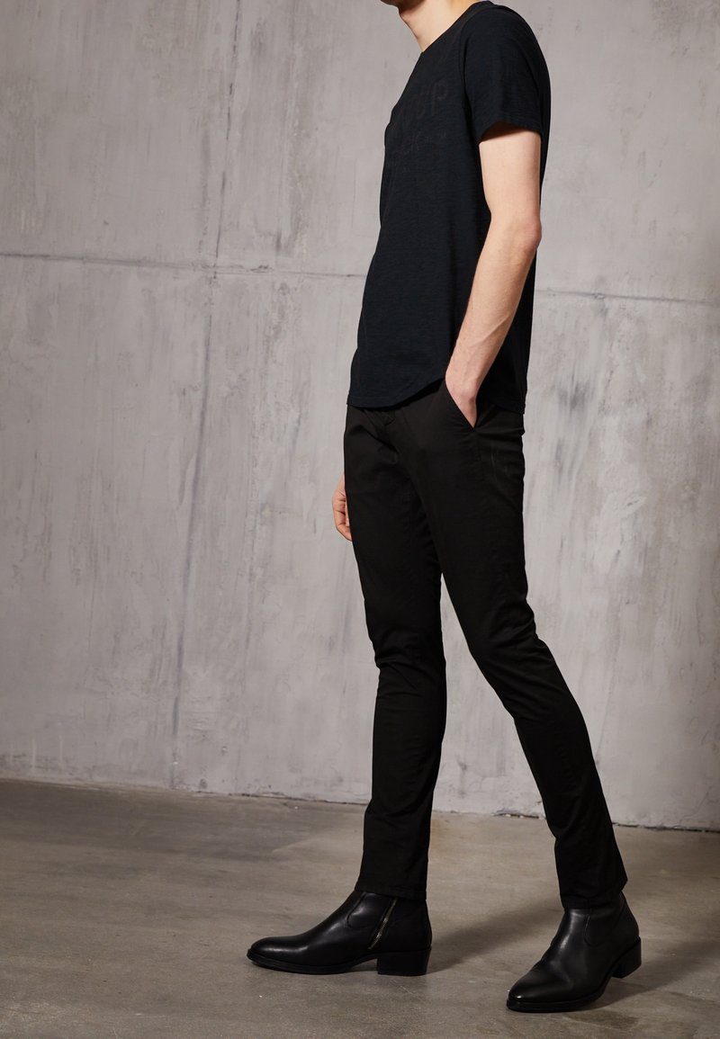 Homme portant un pantalon noir slim, des bottes noires assorties et une chemise noire à manches courtes, debout contre un mur en béton gris.