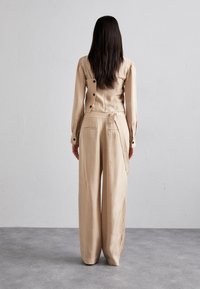 Beige, maßgeschneidertes Outfit mit einem enganliegenden Hemd mit langen Ärmeln und Knöpfen am Rücken sowie hoch taillierten, weiten Hosen. Glatte Textur, minimales Design.
