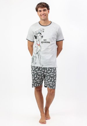 Joven de pie descalzo con un pijama gris estampado de dálmata y el texto "101 Razones" en la camiseta, sonriendo a la cámara.