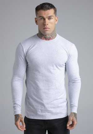 Nyakán és kezén tetoválásokkal rendelkező férfi világosszürke hosszú ujjú ingben és fekete nadrágban, egyszerű szürke háttér előtt áll.