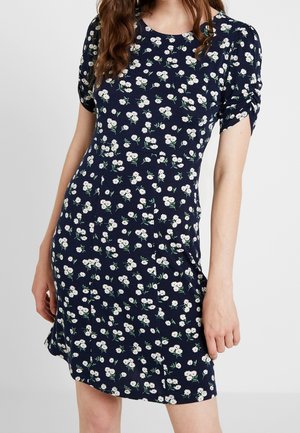 Femme portant une robe bleu marine à manches courtes avec un motif floral de marguerites blanches sur un fond clair uni.